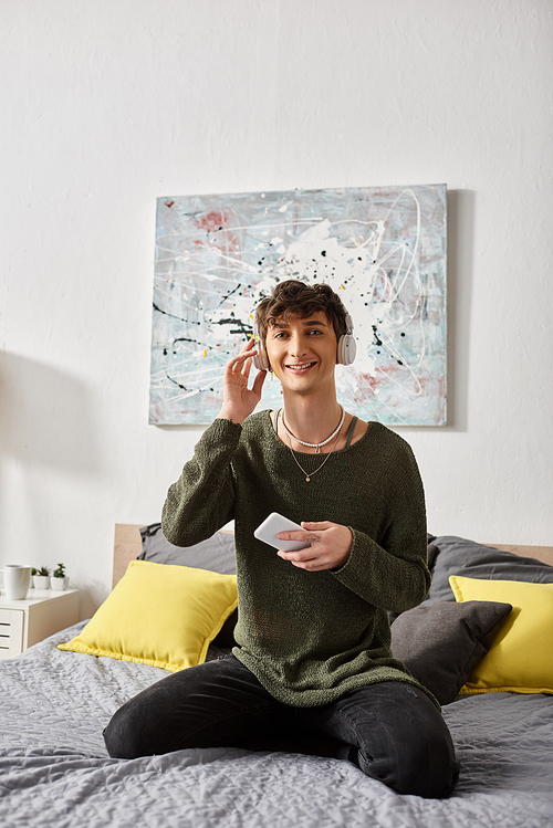 cheerful curly transgender person in wireless headphones holding smartphone and sitting on bed