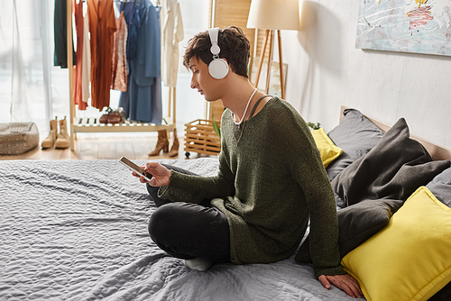 curly transgender person in wireless headphones using smartphone and sitting on bed, social media