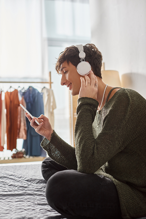 happy curly transgender person in wireless headphones holding smartphone and sitting on bed