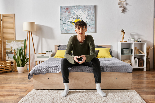 young transgender person in casual attire looking at camera and sitting on bed, modern apartment