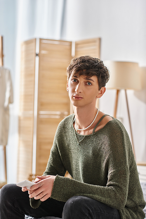 young transgender person in casual attire looking at camera and sitting in modern apartment