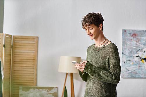 joyful curly transgender blogger using smartphone and standing in bedroom, social media influencer