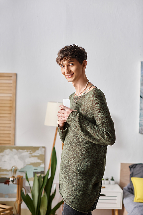 joyful curly transgender blogger using smartphone and standing in bedroom, social media influencer