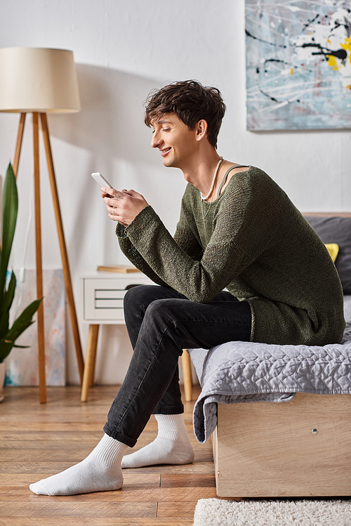 cheerful and curly transgender influencer using smartphone and sitting on bed, social media