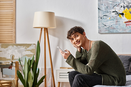 cheerful and curly transgender influencer using smartphone and sitting on bed, social media