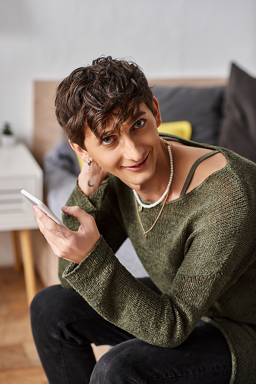 happy and curly transgender influencer using smartphone and sitting on bed, social media