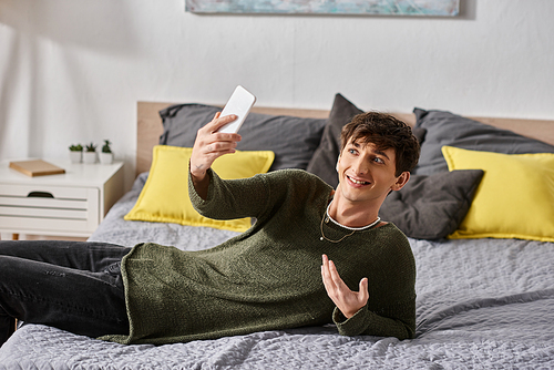 cheerful and curly transgender social media influencer taking selfie on smartphone and lying on bed