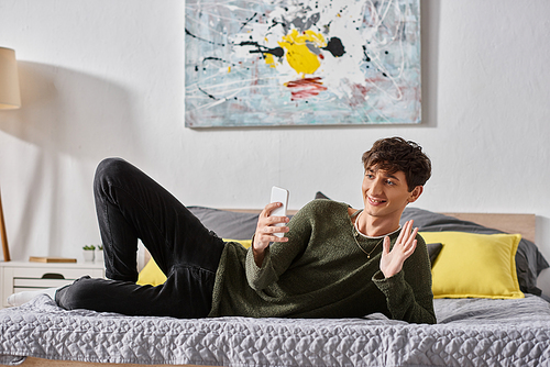 happy and curly transgender social media influencer waving hand during online streaming in bedroom