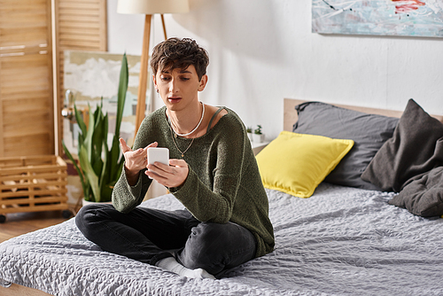 happy and curly transgender influencer using smartphone and sitting on bed, social media