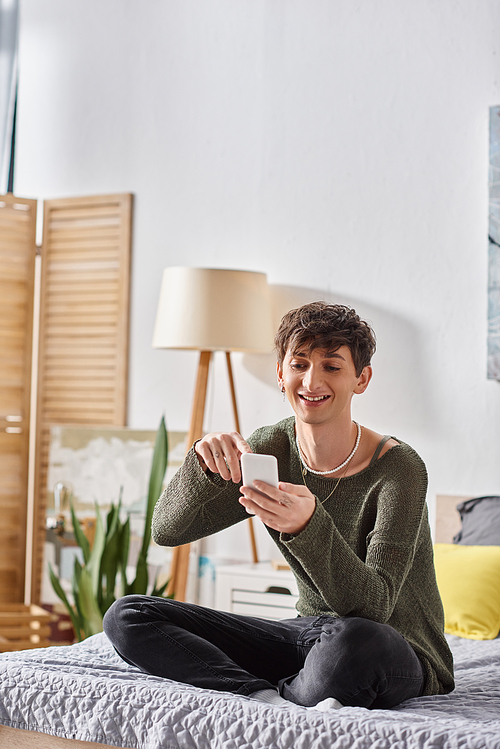 happy and curly transgender influencer using smartphone and sitting on bed, social media