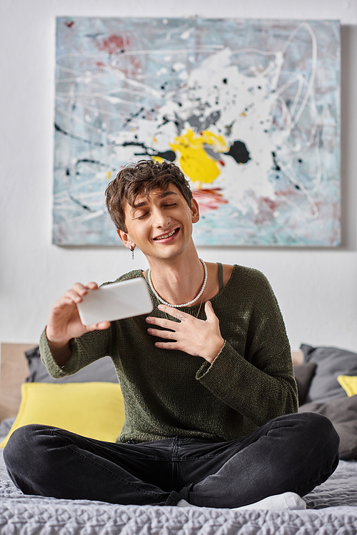 happy curly transgender influencer smiling and using smartphone while sitting on bed, social media