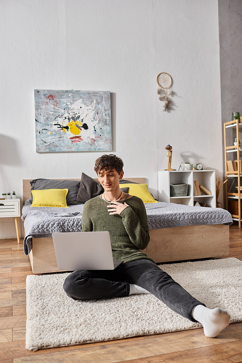 happy and curly transgender influencer using laptop and sitting on carpet near bed, blogger