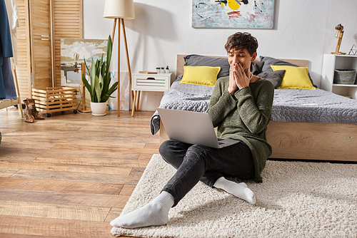 happy curly transgender influencer covering mouth and using laptop while sitting near bed, blogger