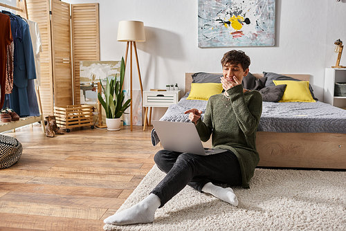 joyful curly transgender influencer covering mouth and using laptop while sitting near bed, blogger