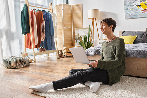 happy and curly transgender influencer using laptop and sitting on carpet near bed, blogger