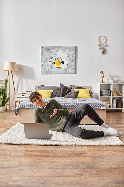 happy and curly transgender influencer using laptop and lying on carpet near bed, social media