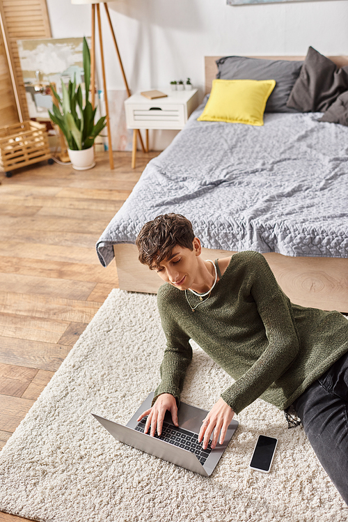 happy curly transgender influencer using laptop and sitting on carpet next to smartphone, blogger