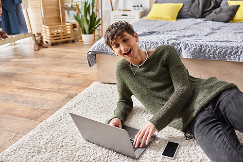 happy curly transgender influencer using laptop and sitting on carpet next to smartphone, blogger