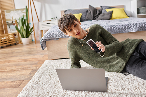 happy transgender influencer using laptop and holding smartphone while sitting on carpet, blogger