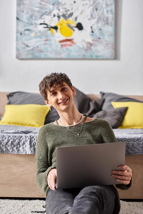 positive and curly transgender influencer using laptop and sitting next to bed, social media