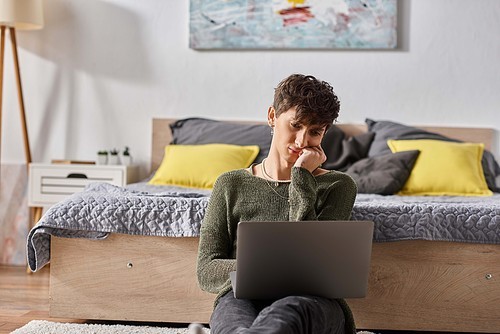 bored and curly transgender influencer using laptop and sitting next to bed, social media