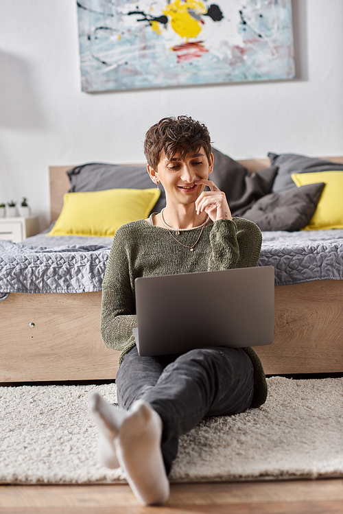 positive and curly transgender influencer using laptop and sitting next to bed, social media