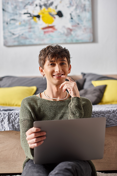 pleased and curly transgender influencer using laptop and sitting next to bed, social media