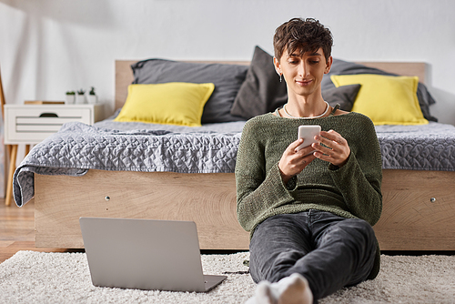 curly transgender blogger using smartphone next to laptop while sitting on carpet, social media