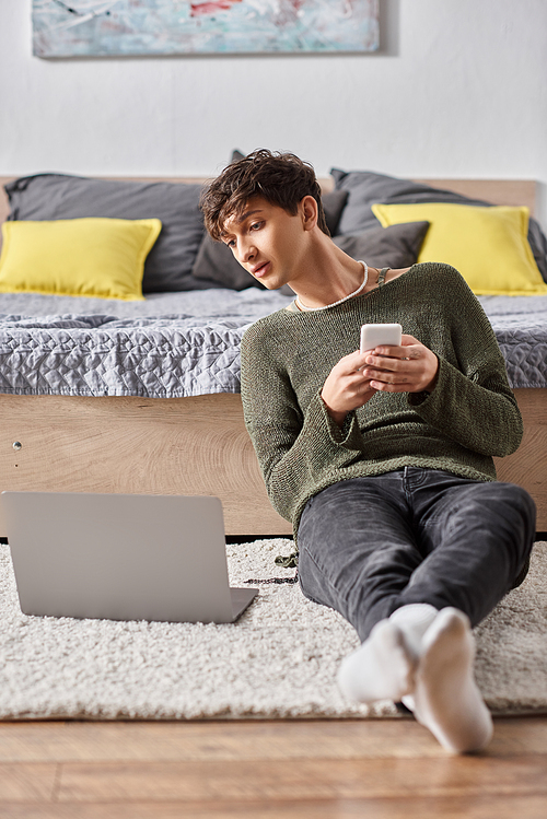 transgender blogger using smartphone and looking at laptop while sitting on carpet, social media