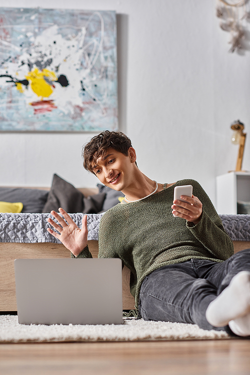 curly transgender blogger holding smartphone and waving hand at laptop while sitting on carpet