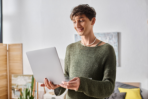 transgender blogger with curly hair using laptop while standing in modern apartment, influencer