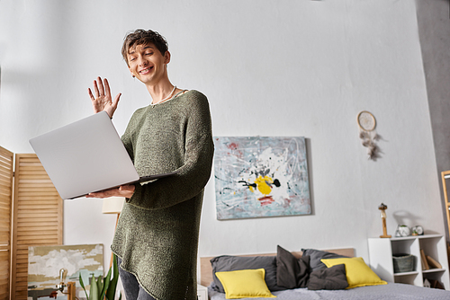 transgender blogger having video call on laptop and standing in modern apartment, happy influencer