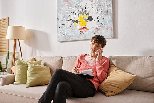 transgender blogger in pink sweater talking on smartphone and writing in notebook, sitting on sofa