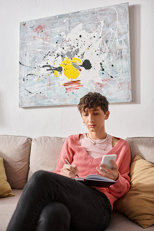 transgender blogger in pink sweater holding smartphone and writing in notebook, sitting on sofa