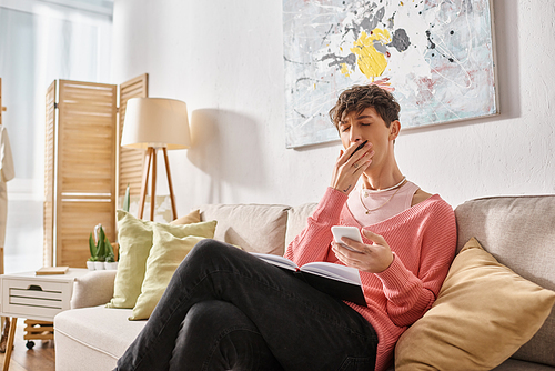 tired transgender blogger in pink sweater holding smartphone and yawning while sitting on sofa