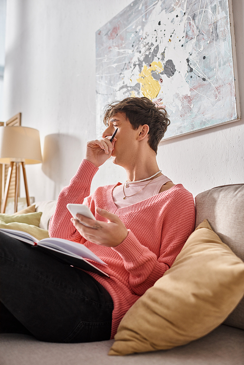 tired transgender blogger in pink sweater holding smartphone and yawning while sitting on sofa