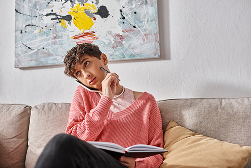 transgender person in pink sweater talking on smartphone and writing in notebook, sitting on sofa