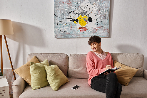 happy transgender blogger in pink sweater writing in notebook and looking at smartphone on sofa