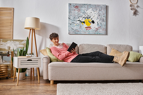 happy transgender person in pink sweater writing in notebook and sitting on sofa next to smartphone