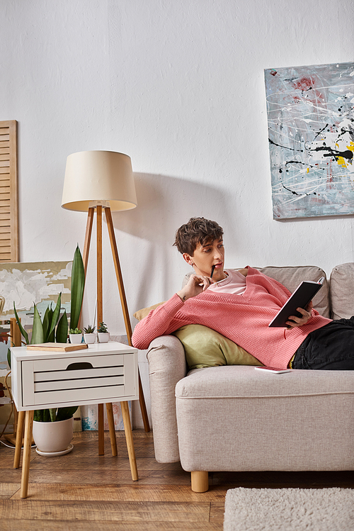 pensive transgender person in pink sweater writing in notebook and sitting on sofa with smartphone