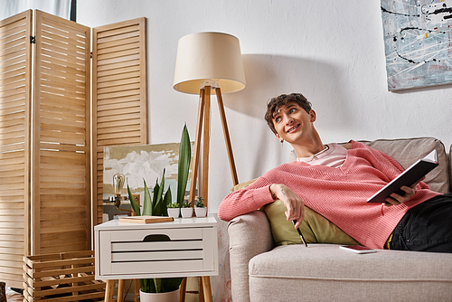 cheerful transgender person in pink sweater holding notebook and sitting on sofa next to smartphone