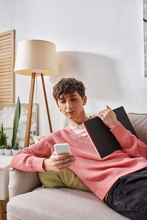 curly transgender person in pink sweater holding notebook, using smartphone and sitting on sofa