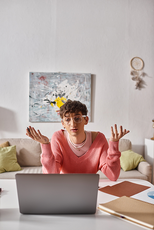 young transgender freelancer in pink sweater gesturing while looking at laptop on desk, video call