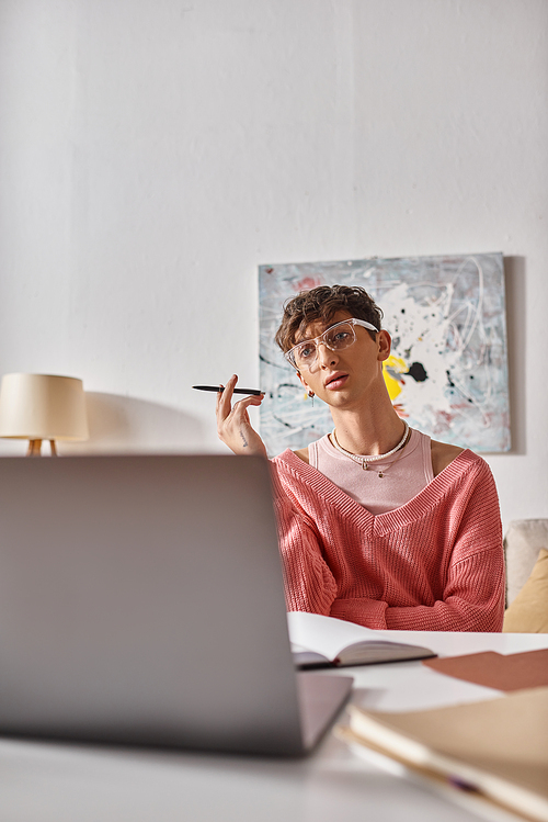 young transgender freelancer in pink sweater gesturing while looking at laptop on desk, video call