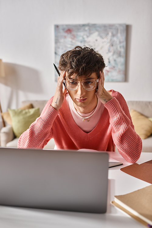 young transgender freelancer in pink sweater and eyeglasses looking at laptop on desk, remote work