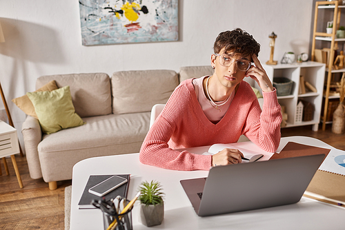 pensive transgender freelancer in pink sweater and eyeglasses looking away while working remotely