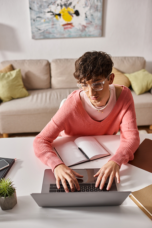 young androgynous freelancer in pink sweater and eyeglasses typing on laptop while working remotely