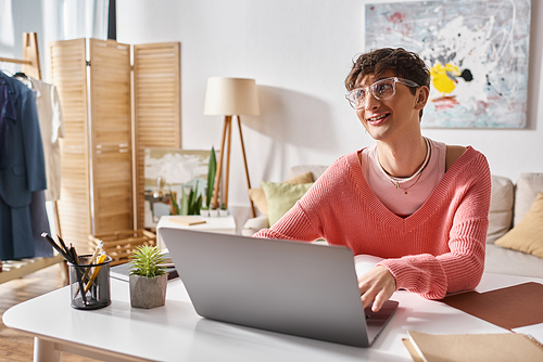 happy androgynous freelancer in pink sweater and eyeglasses typing on laptop while working remotely