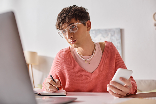 curly androgynous freelancer in pink sweater and eyeglasses holding smartphone and using laptop