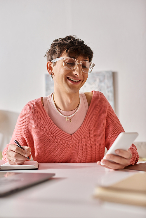 happy androgynous blogger in pink sweater and eyeglasses using smartphone and taking notes